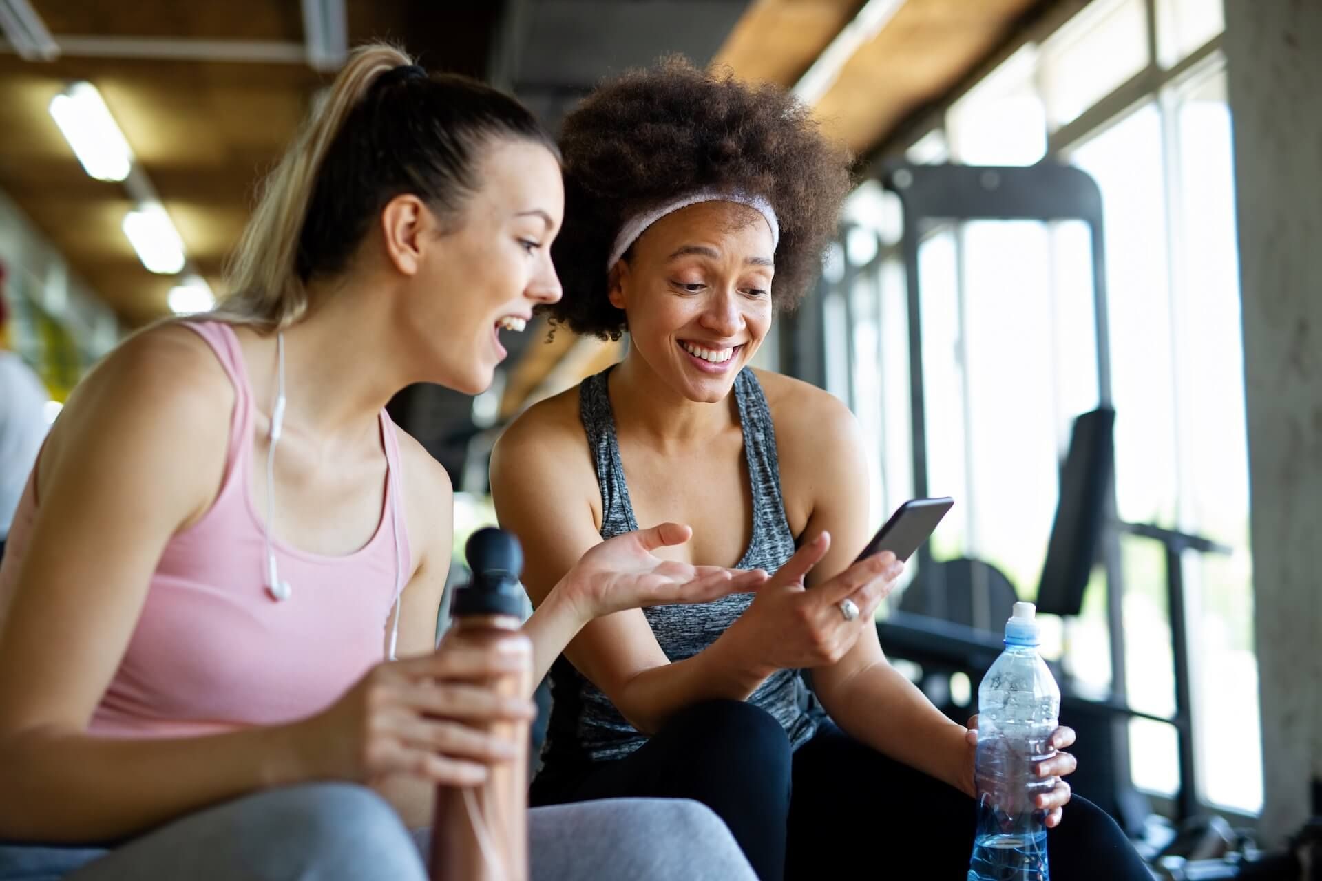Two women resting with water bottles after a workout at the gym