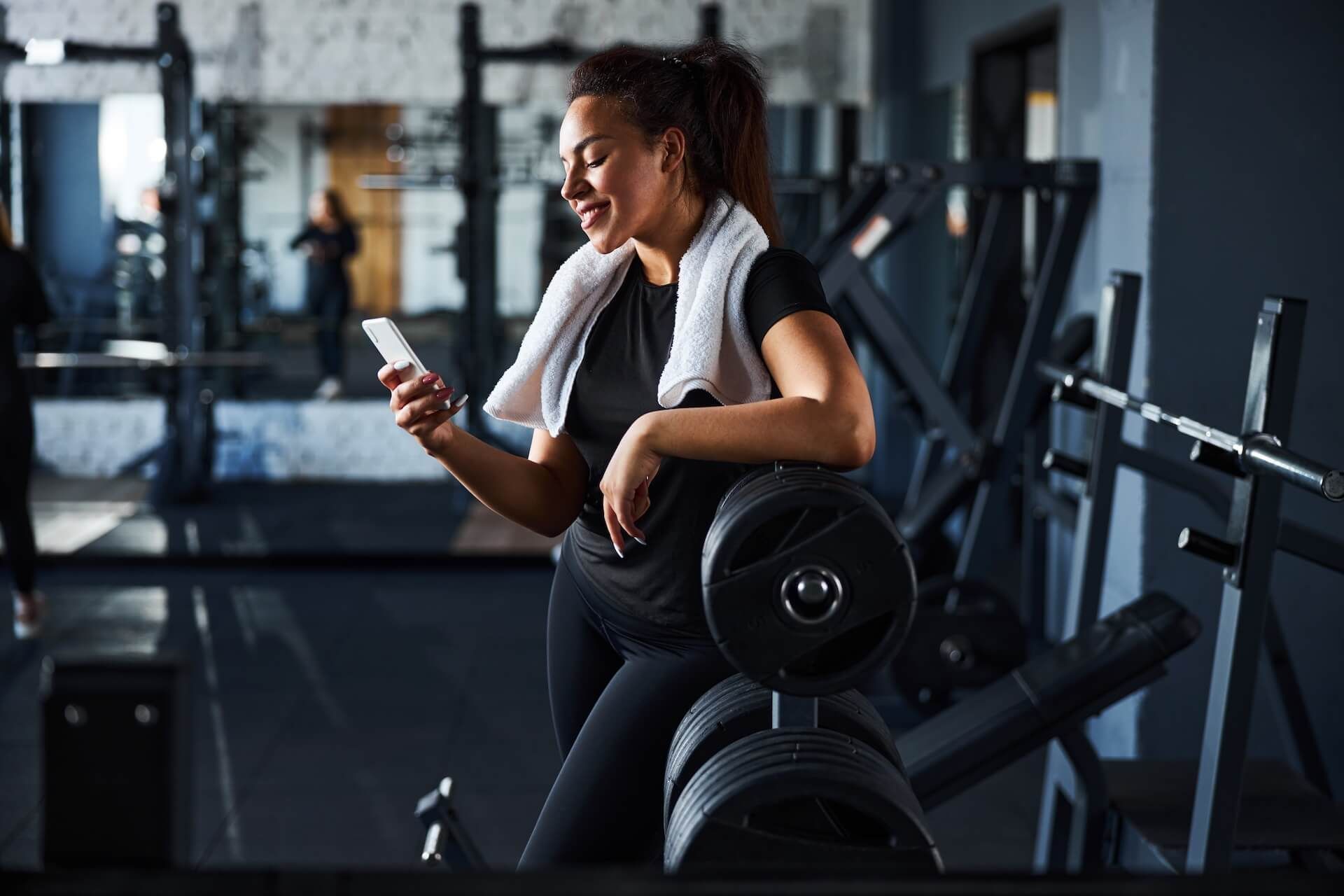Woman resting with a towel, checking her phone in a gym beside weight machines and dumbbells.