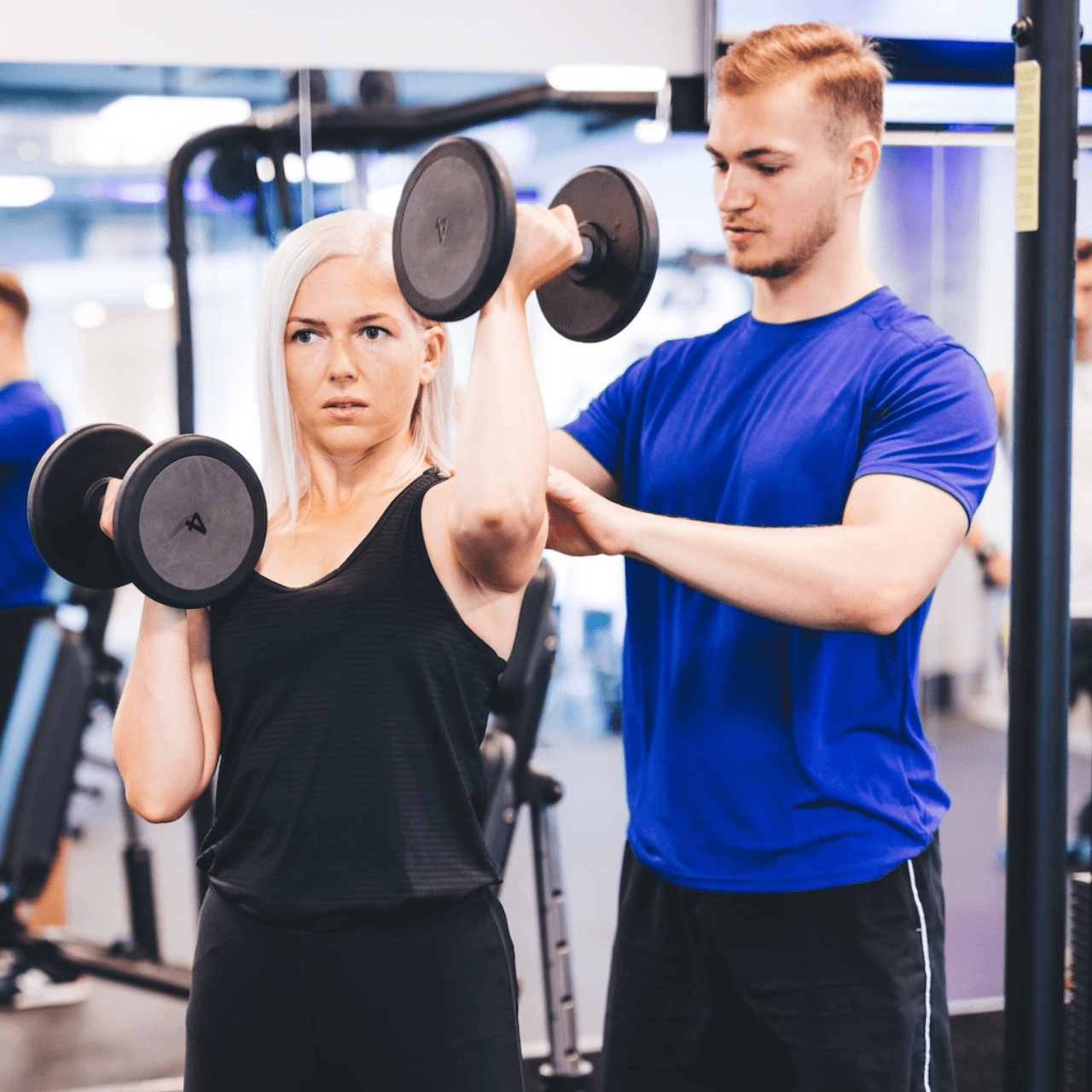 Two people doing dumbbell shoulder presses in a gym, with a trainer spotting from behind.