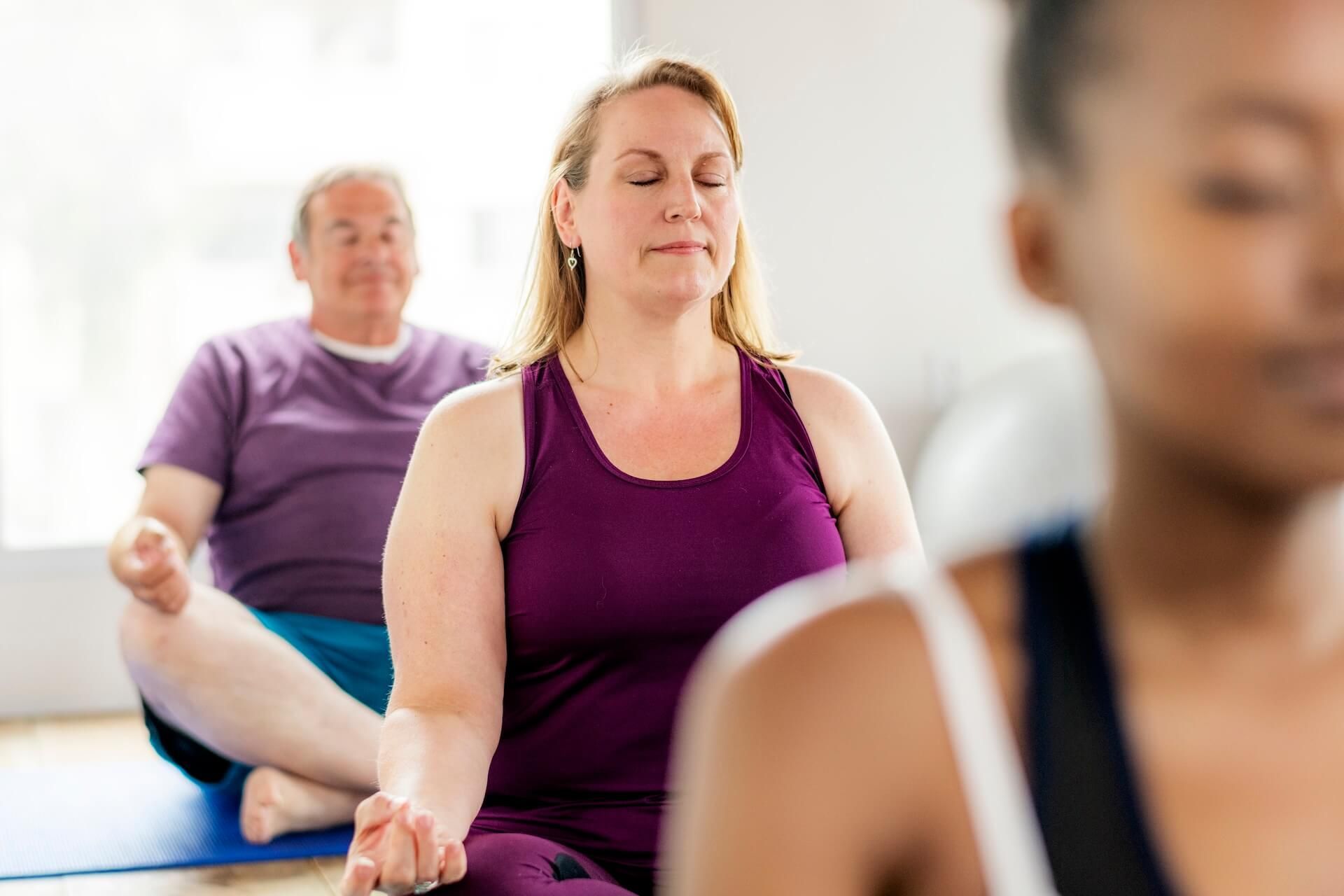 People meditating in a bright yoga class, one woman in a purple top in focus.
