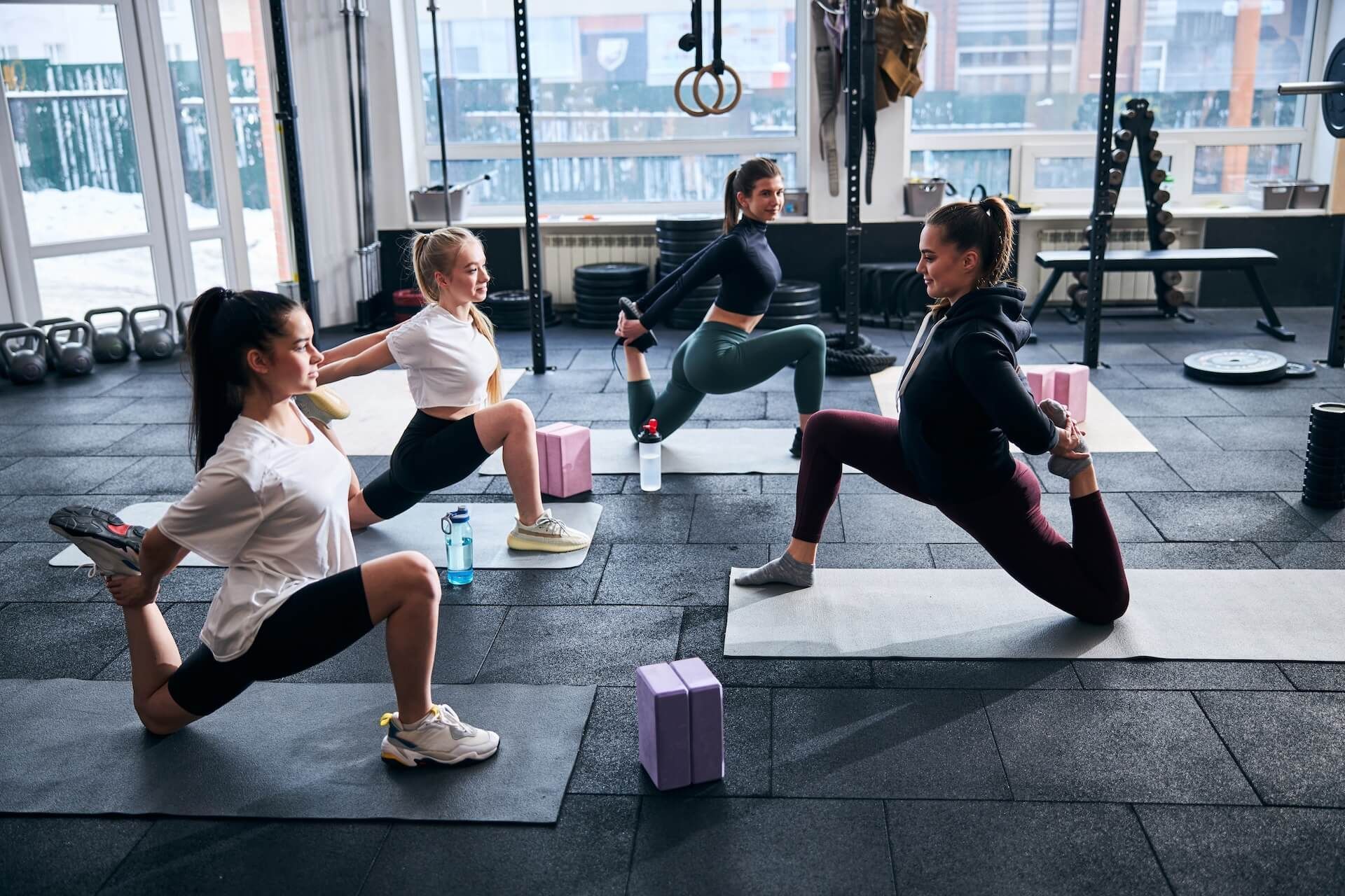 Fitness class stretching on mats in a bright gym with kettlebells and large windows.