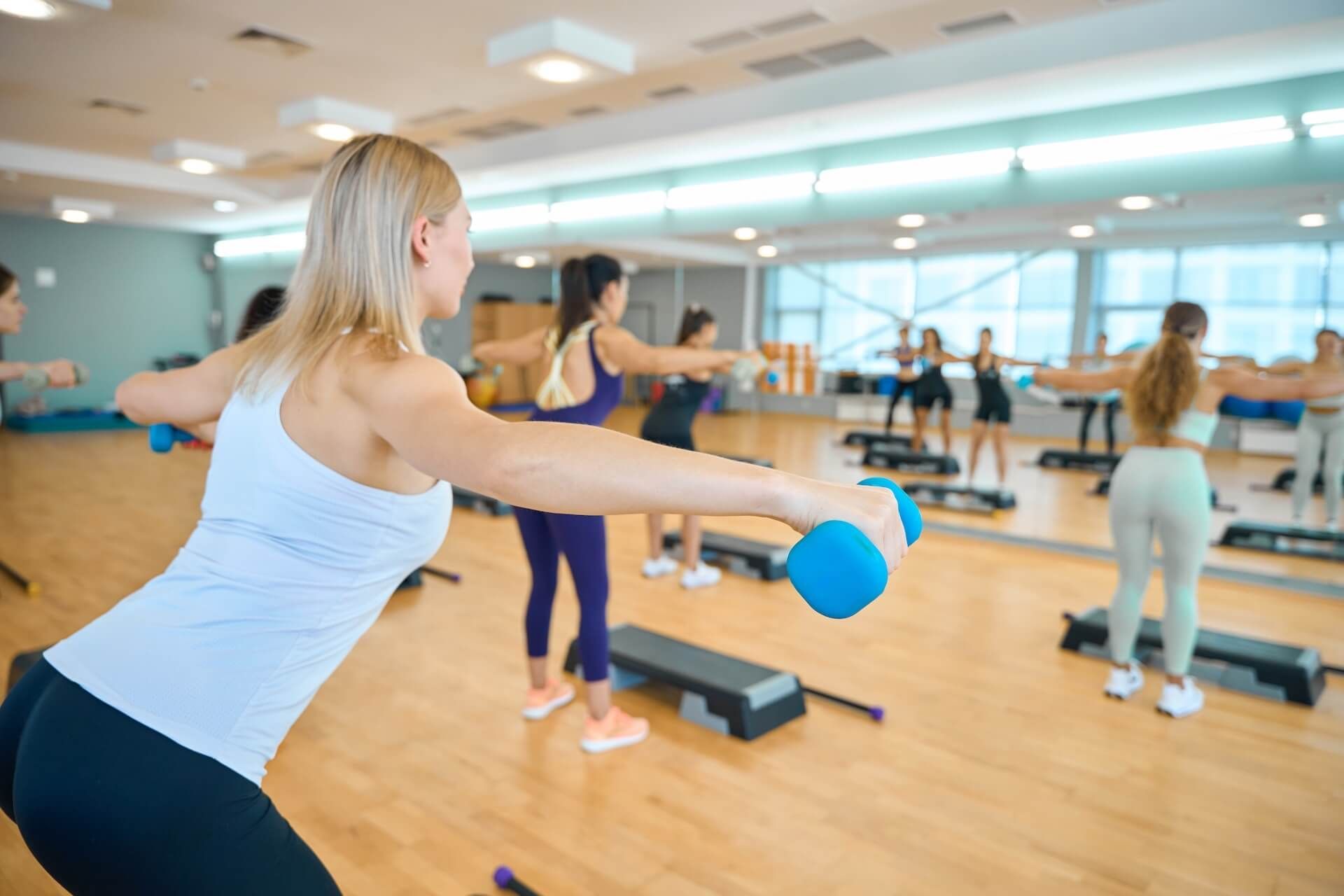 Group fitness class in a mirrored studio, with people lifting blue dumbbells and using step platforms.