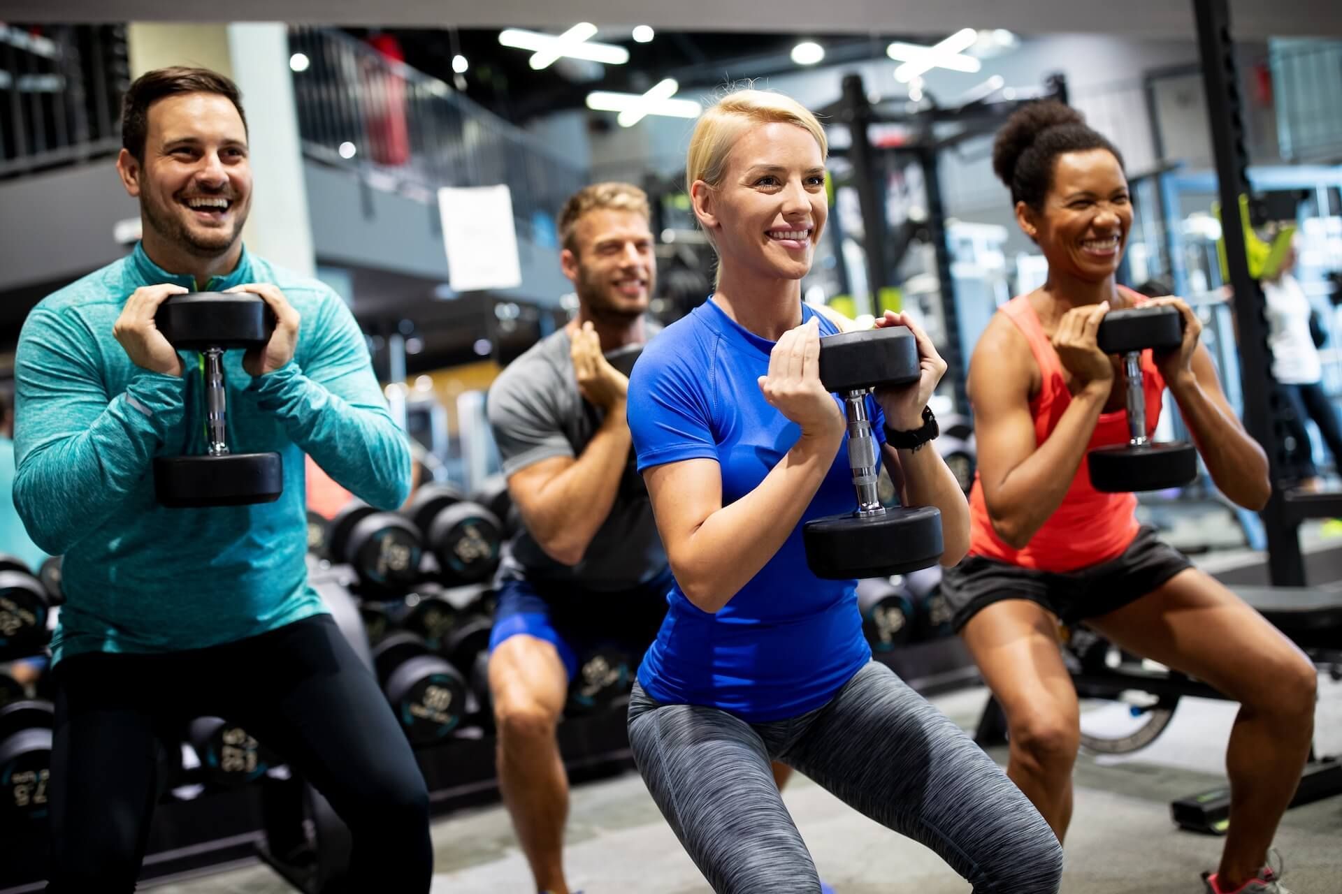 People doing dumbbell squats in a gym, smiling and exercising together