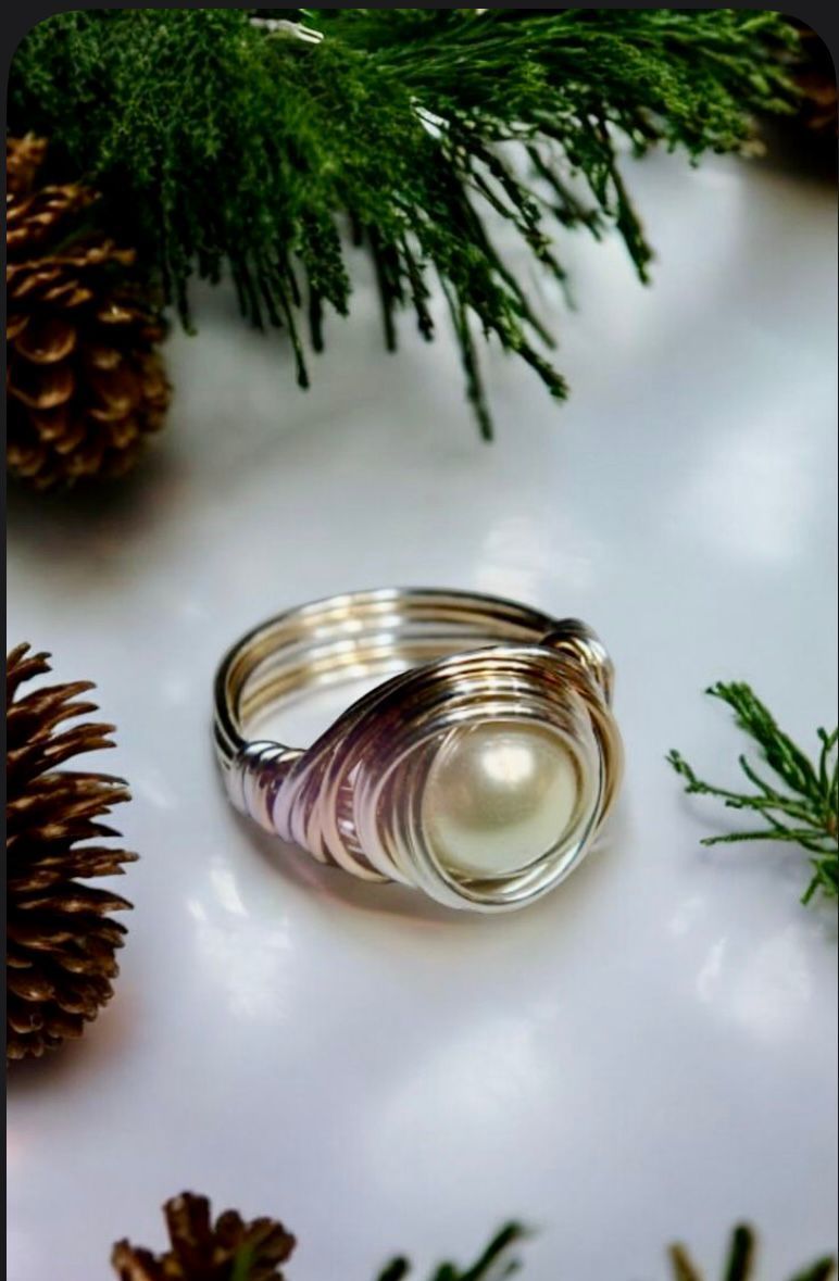 Anillos dorados apilados con un centro de perla pálida sobre tela blanca, enmarcados por ramitas de pino y una piña.