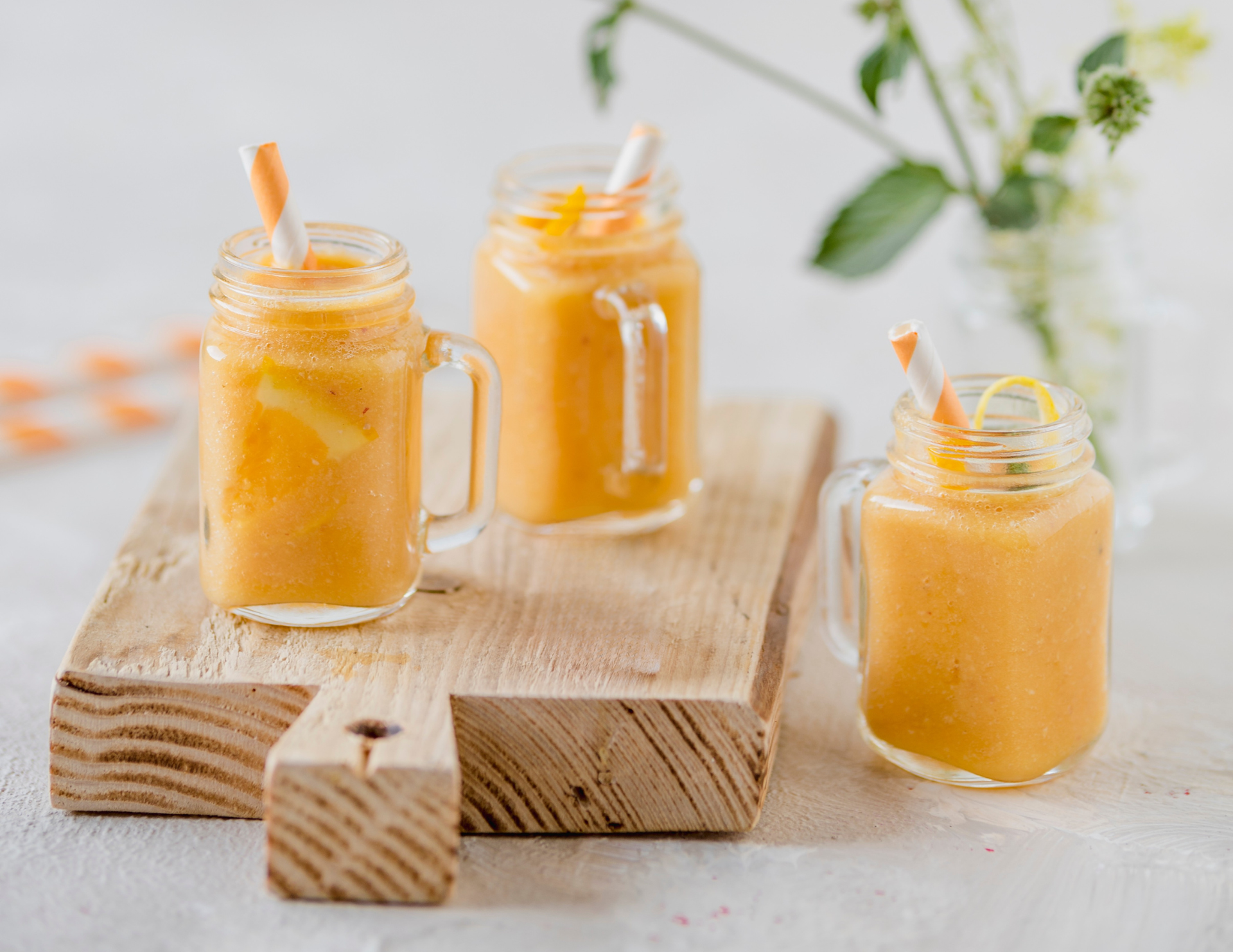 Three glass mugs, filled with Orange Creamsicle Smoothies sit on a cutting board on a white table with folliage in the back