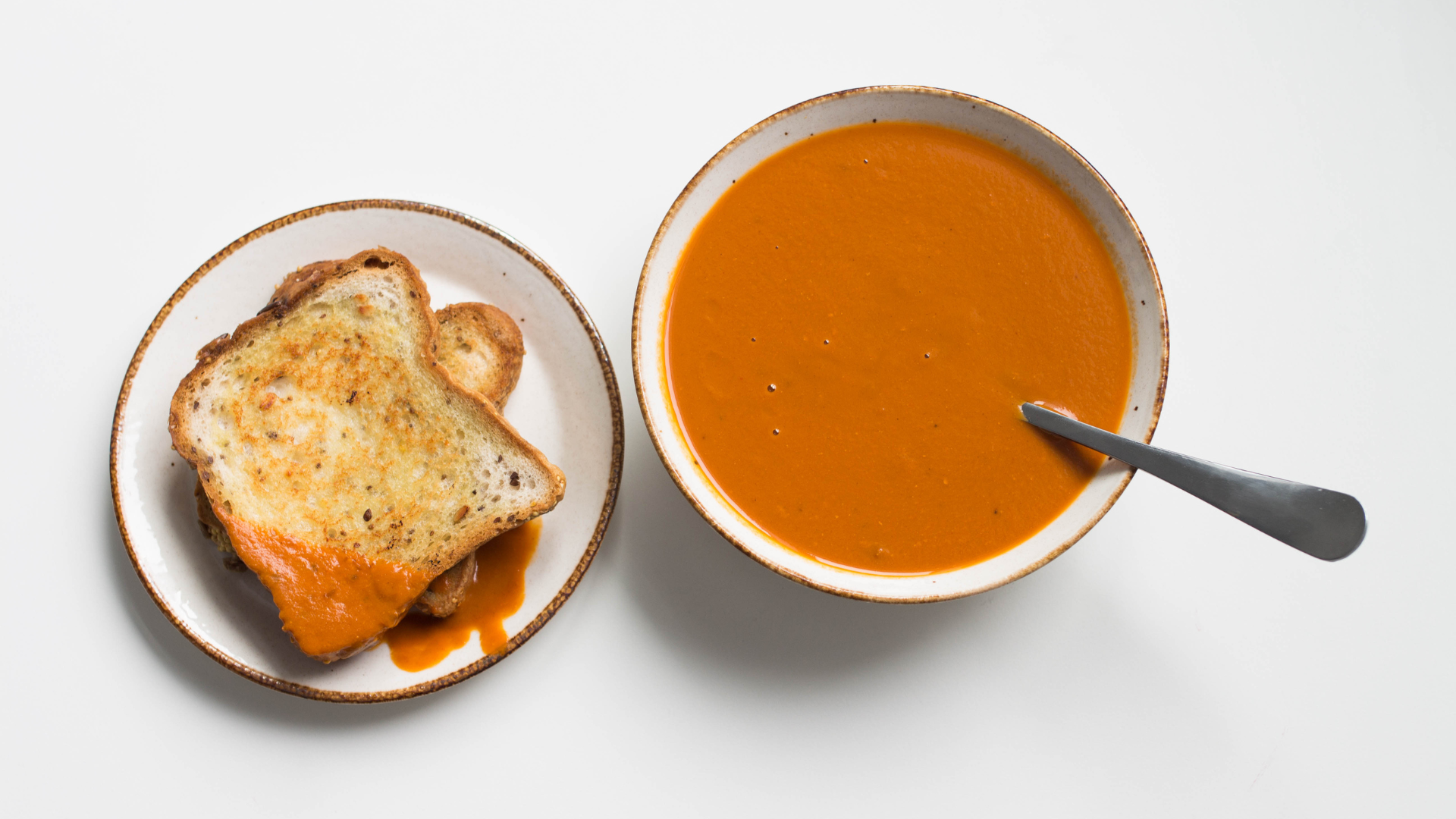 A bowl of Tomato soup with a spoon sticking out. Next to it is a plate with two pieces of toast, one of them has been dunked