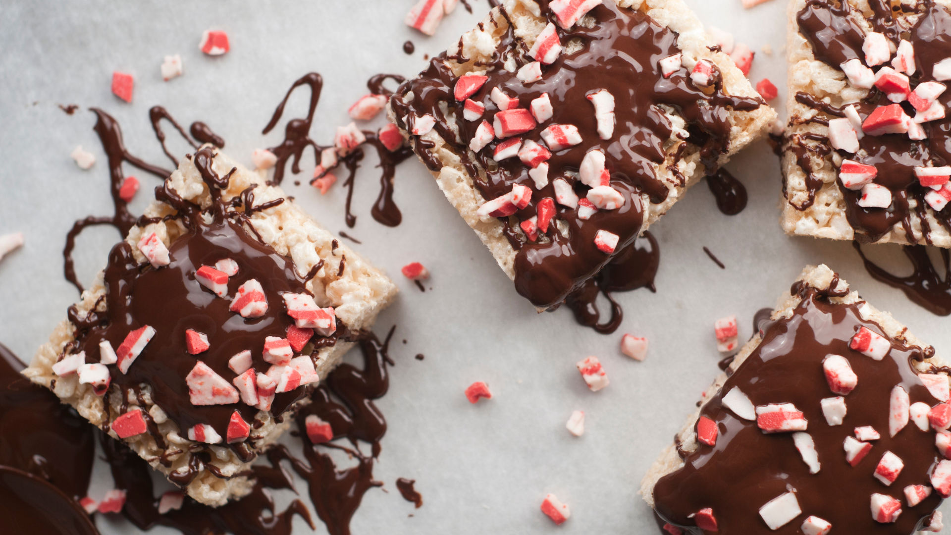Rice Krispie treats sit on parchment paper, covered in a chocolate sauce and bits of candy cane garnish the top