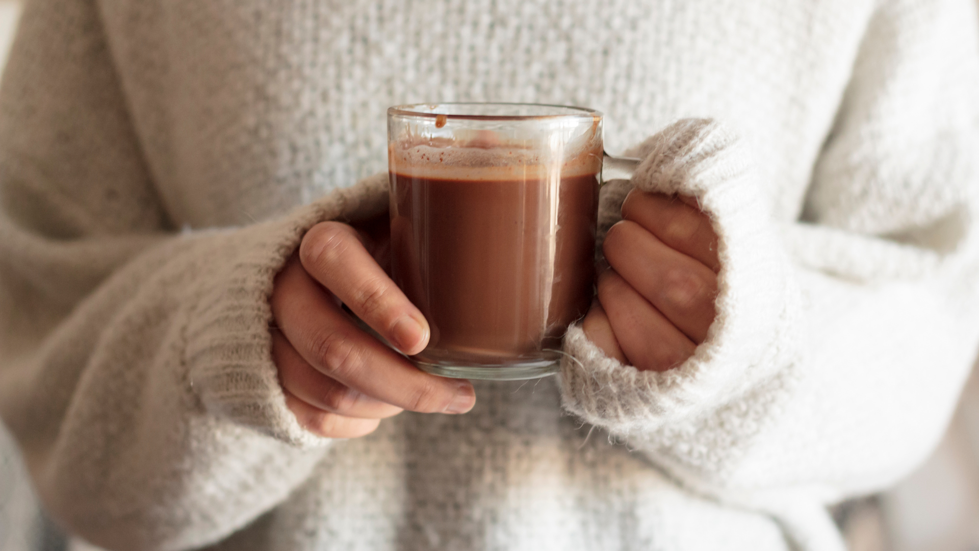 A small glass mug filled with hot chocolate is held by a person wearing a large fluffy white sweater