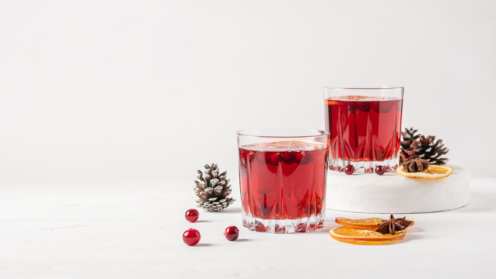 Two glasses containing a red drink sit against a white background surrounded by pinecones and orange slices.