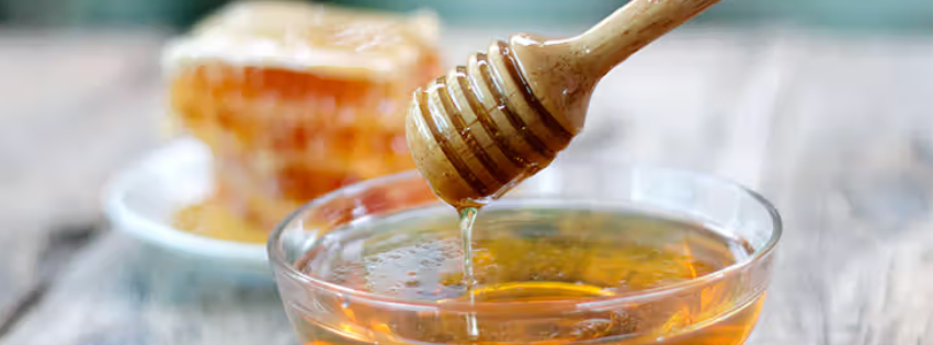 A honey dipper is held above a bowl of honey, with some of the honey dripping off the dipper into the bowl