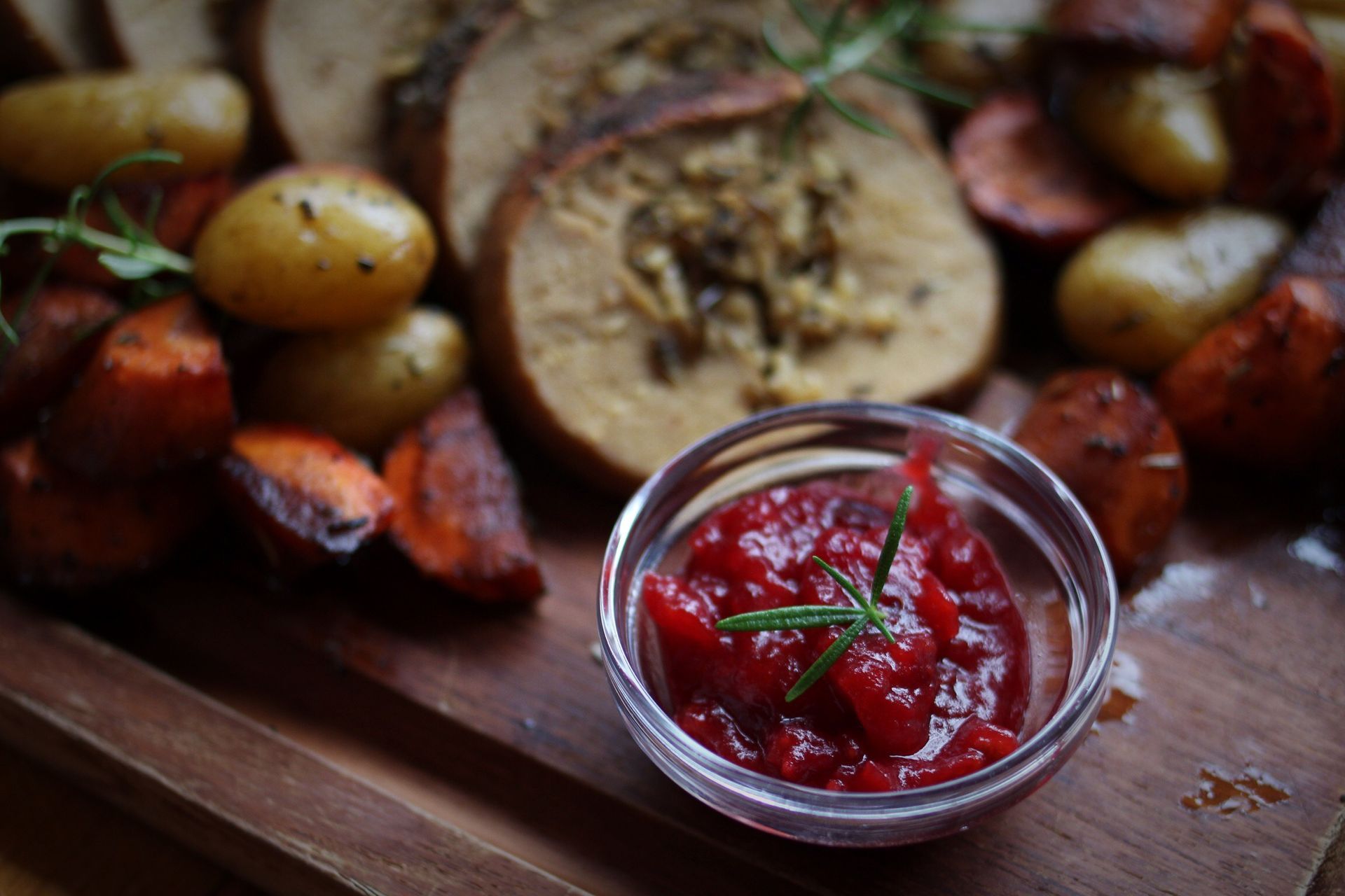 A small glass bowl of cranberry sauce sits on a cutting board in front of bread and raw small potatoes