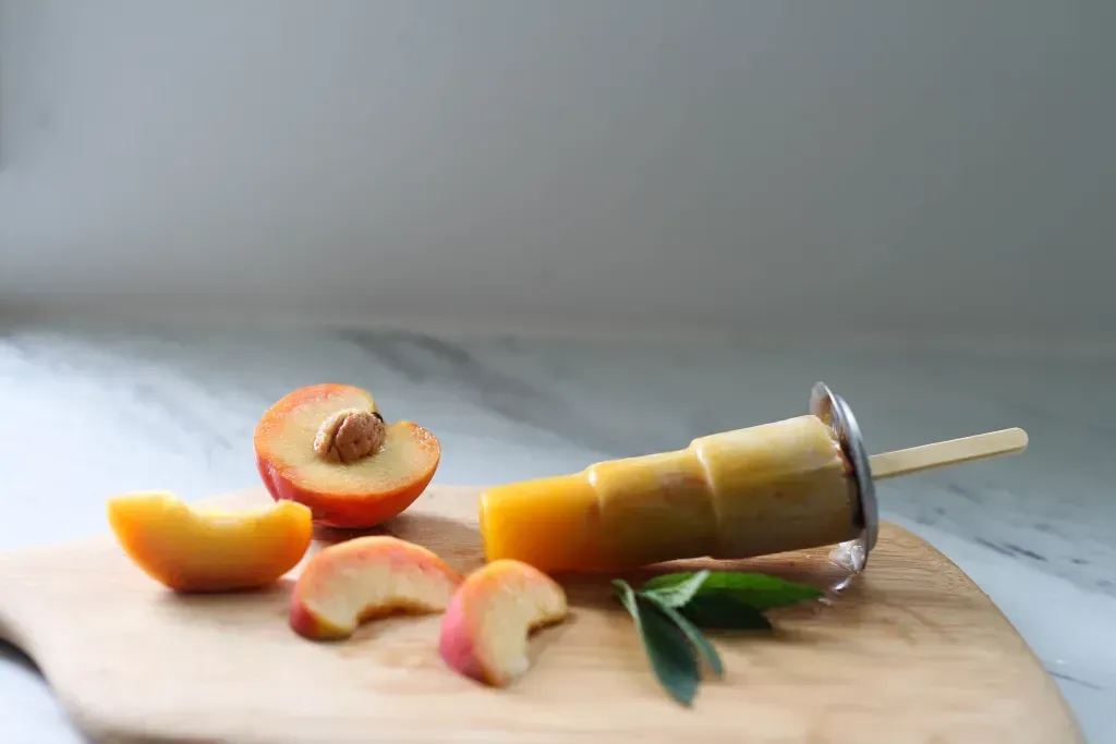 An orange popsicle sits on a cutting board along with peach slices