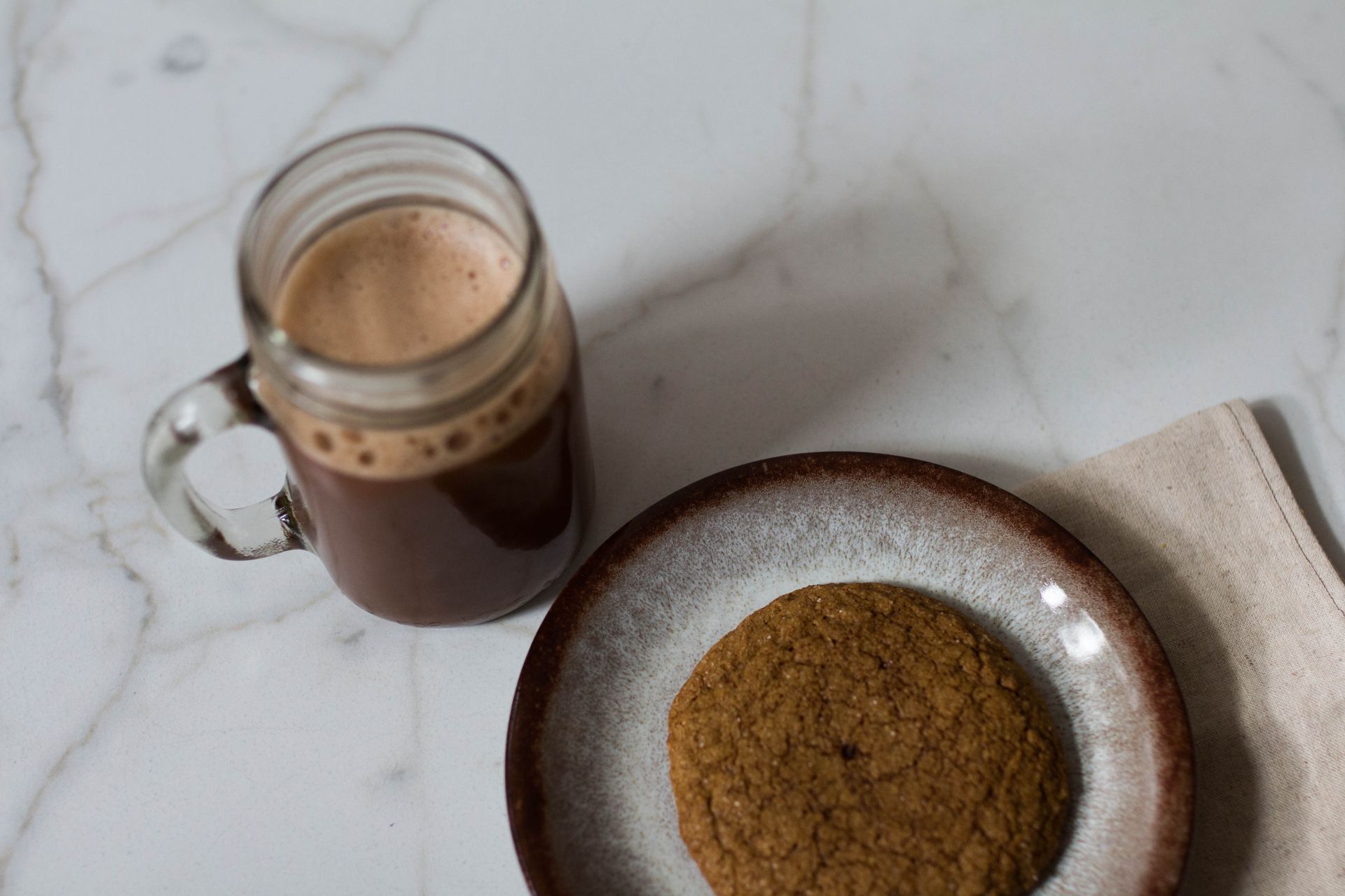 A ginger cookie sits on a plate next to a glass mug of hot chocolate. Both sit on a white table with a black design.