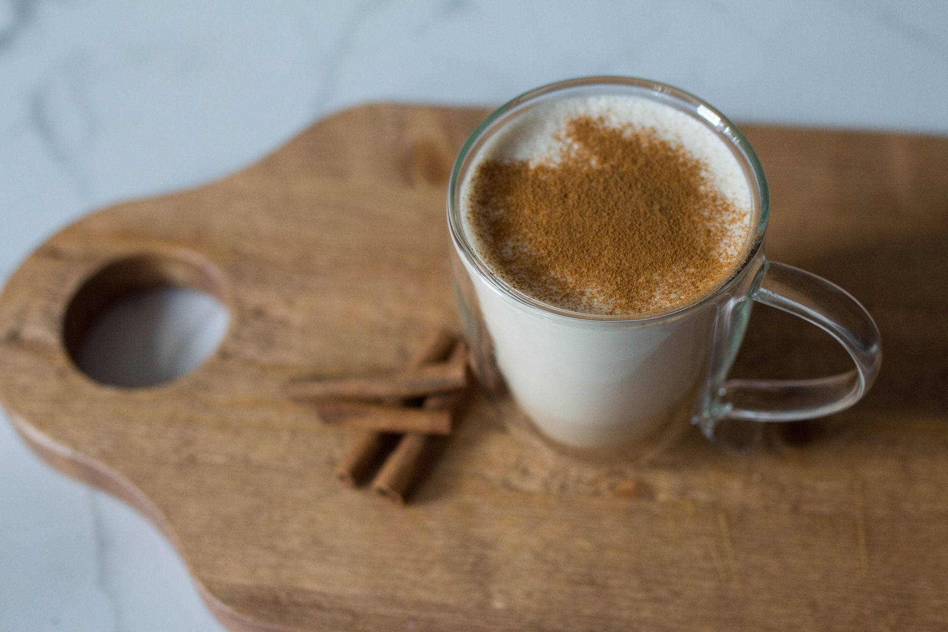 A white beverage sits in a glass mug with cinnamon covering the top. Underneath is a cutting board and cinnamon sticks.