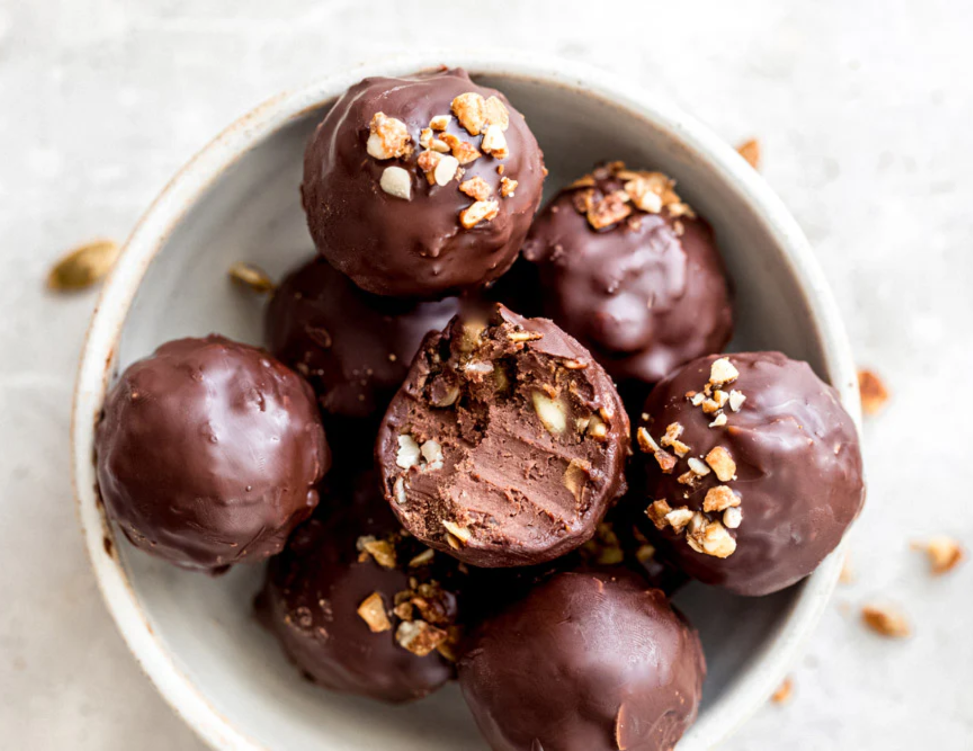 A bowl of chocolate peanut butter collagen truffles with nuts garnishing them sits on a white table with crumbs surrounding