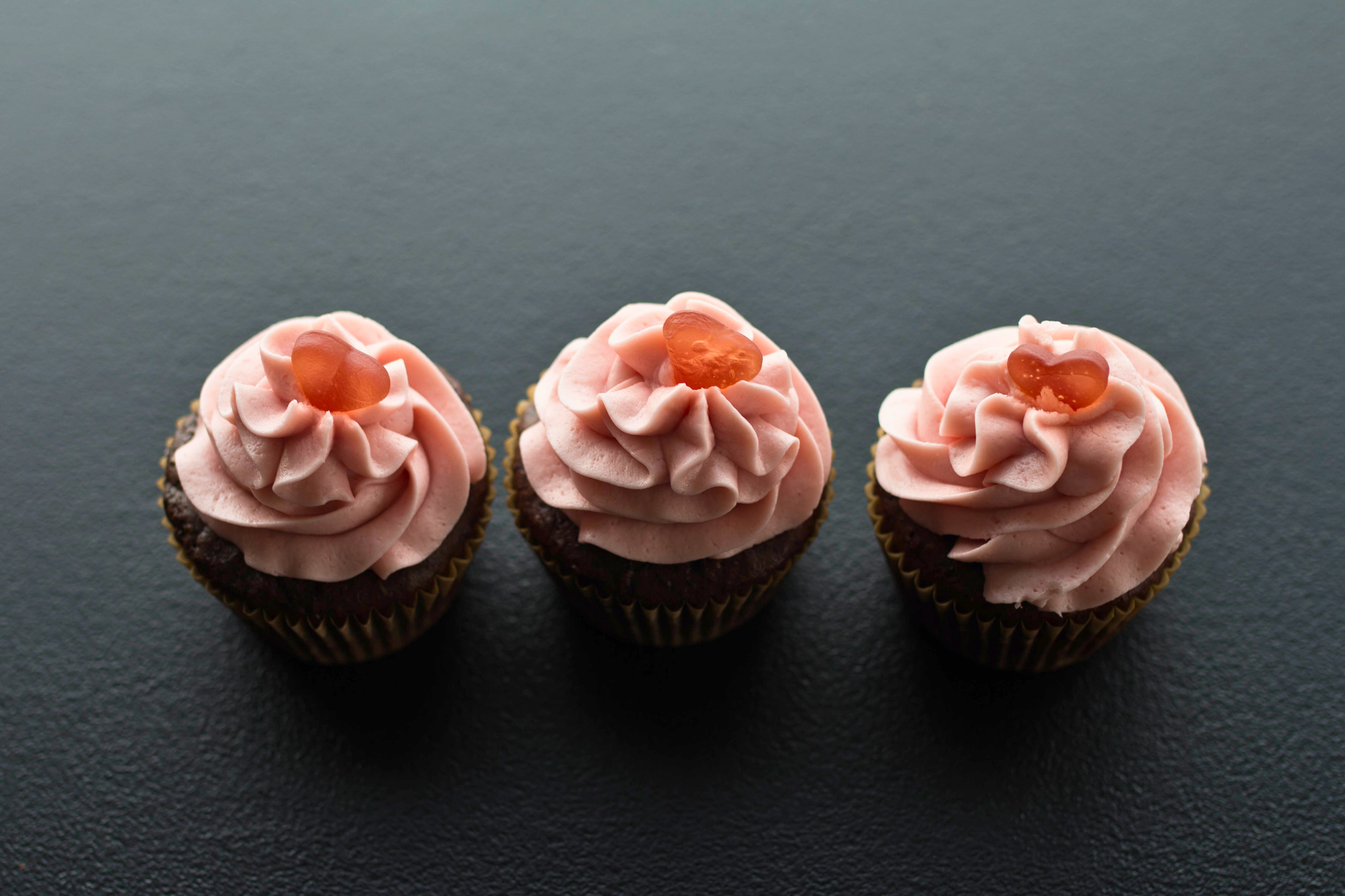 3 cupcakes with pink frosting sit on a table