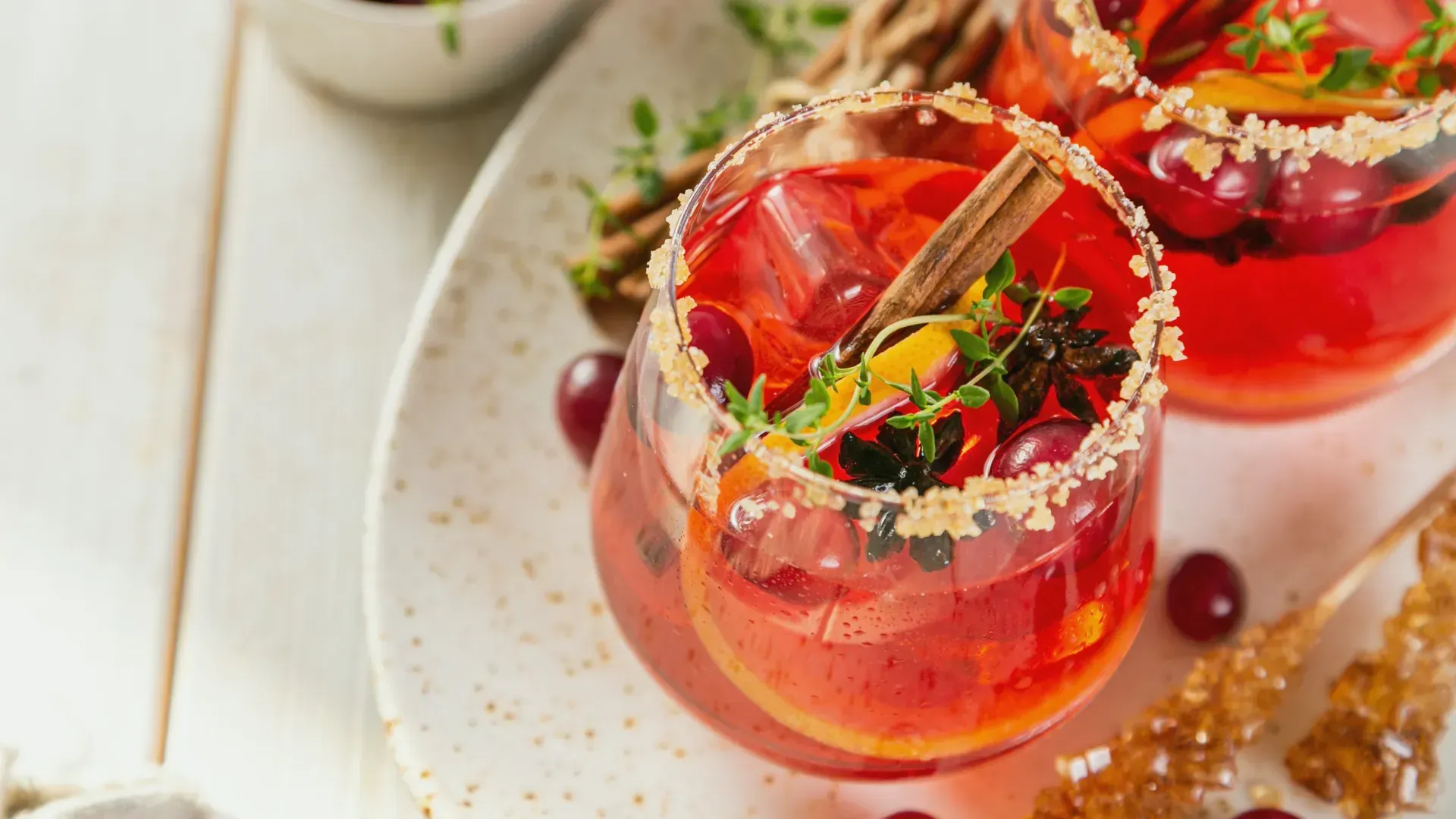 A glass of a red colored drink that is garnished with greens, red grapes and cinnamon sticks. It sits on a plate.