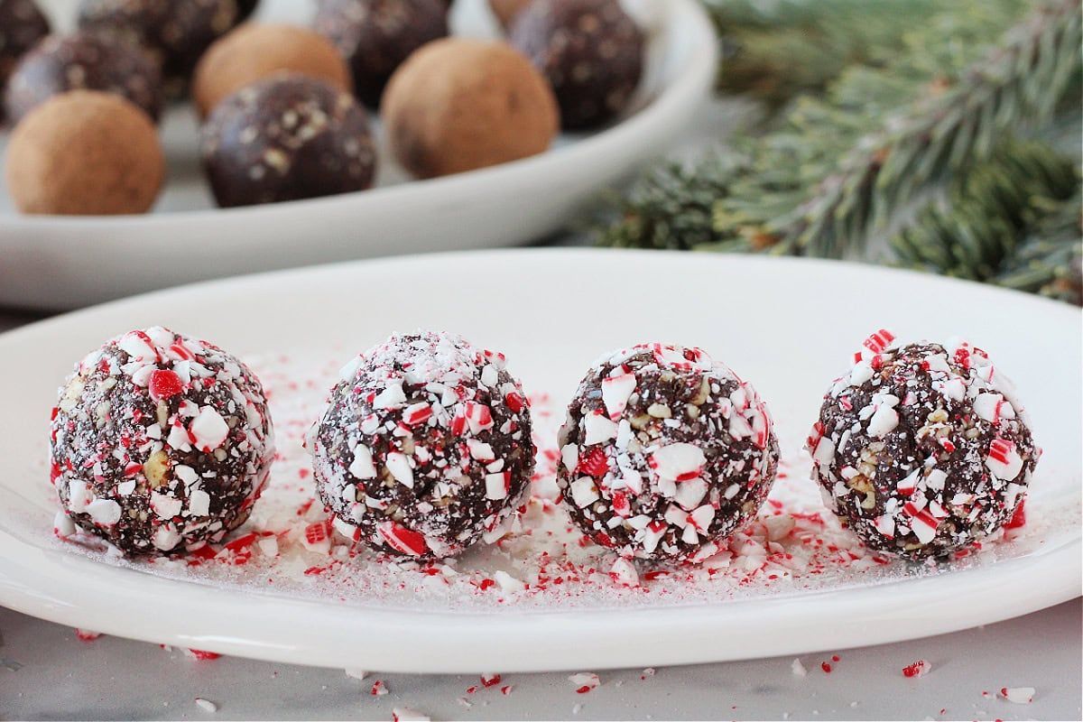 4 chocolate truffles coated with candy cane bits sit on a white plate, other truffles can be seen in the background