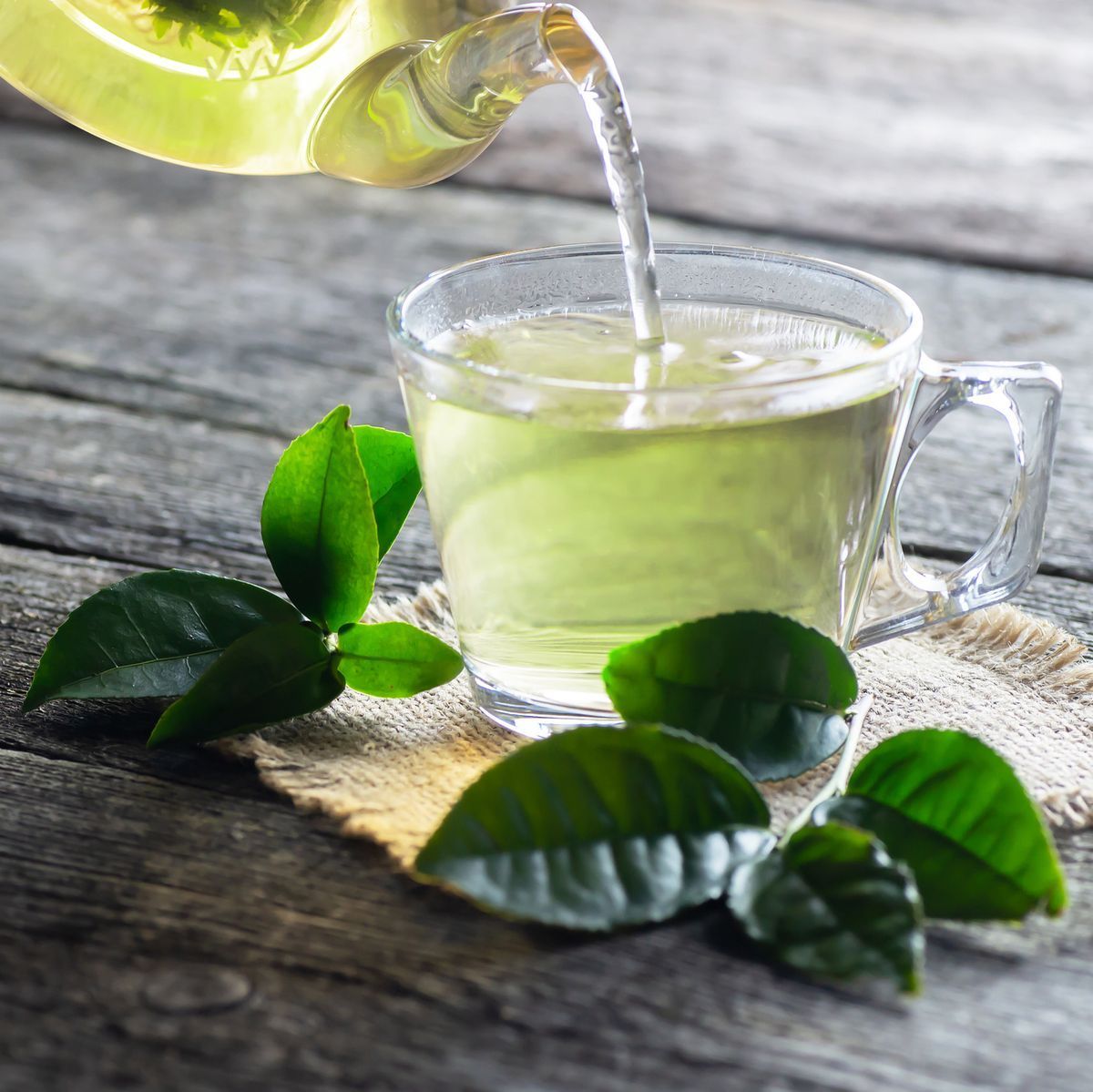 A glass of green tea sits on a wooden table surrounded by green leaves