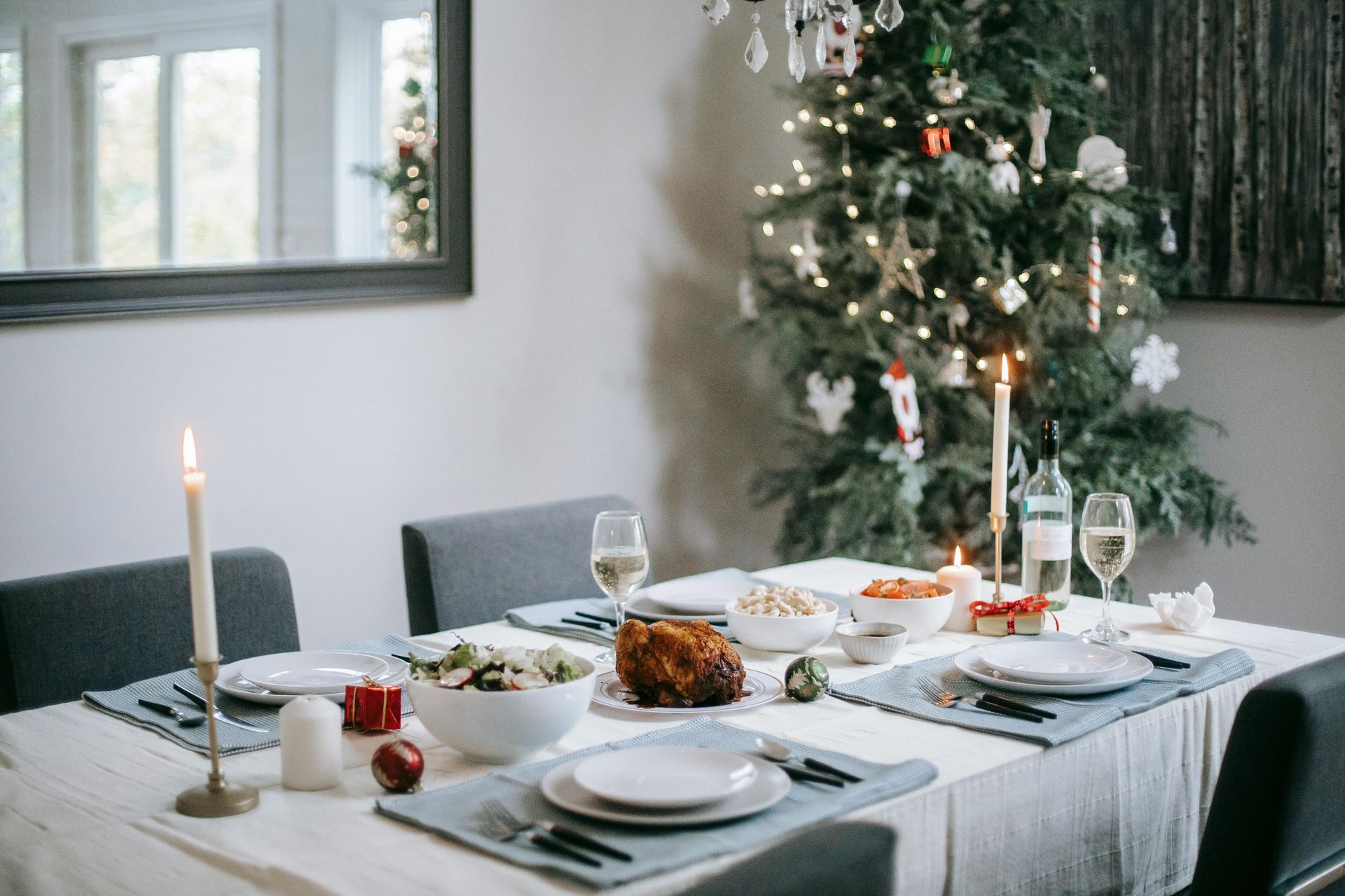 A holiday dinner table setup with dishes, plates, utensils and candles decorating the table. A Christmas tree in the back