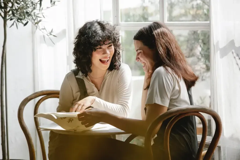 Two women sit at a table and laugh while reading a book