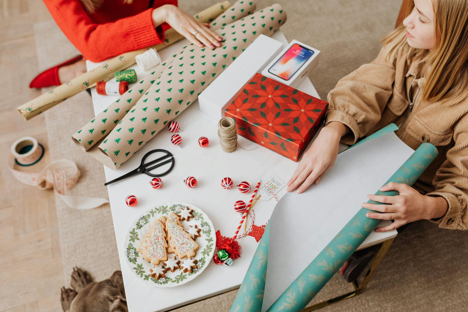 A table is filled with wrapping paper, holiday treats and wrapping supplies. Two women sit at the table.