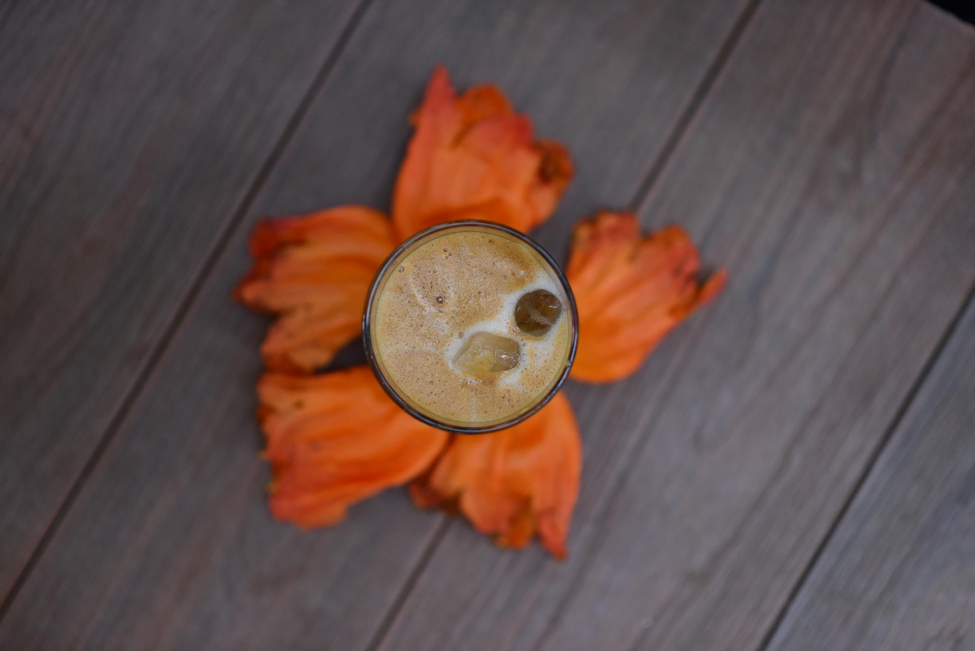 Orange flower petals surround a glass filled with a latte drink, they both sit on a wooden deck with shadows
