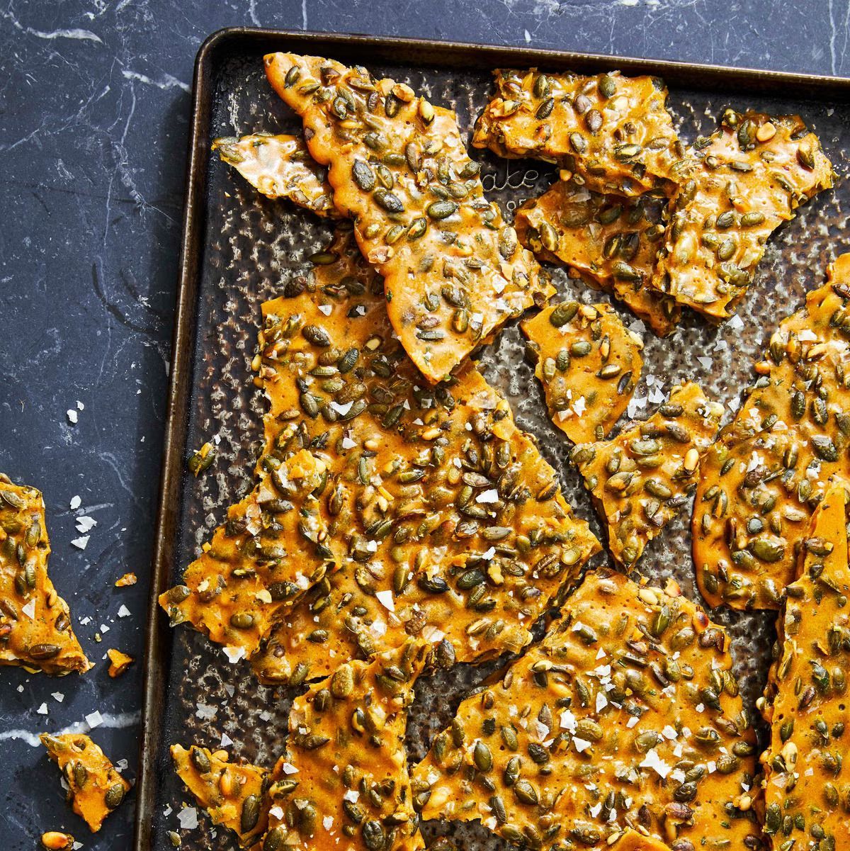 Rosemary-Pumpkin Seed bark sits on a black tray and is cracked up, almost ready to eat. Sits on a dark blue table