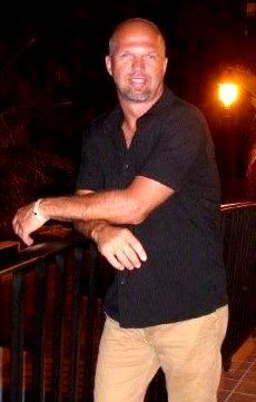A bald man with a beard is standing with his arms crossed in a dark room.