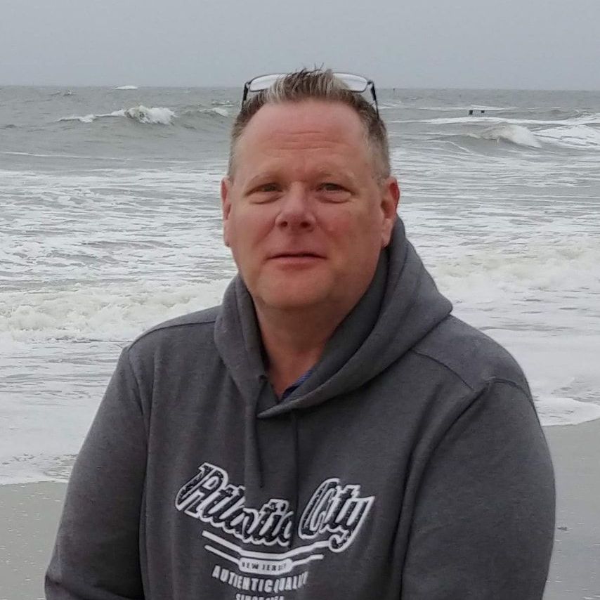 A man is standing on a beach wearing a hoodie and sunglasses.