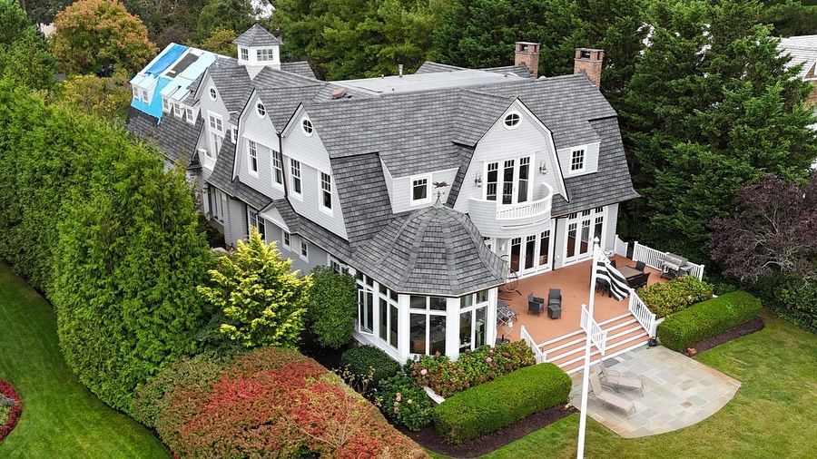 Large gray multi-story house with a complex roof and surrounding greenery.
