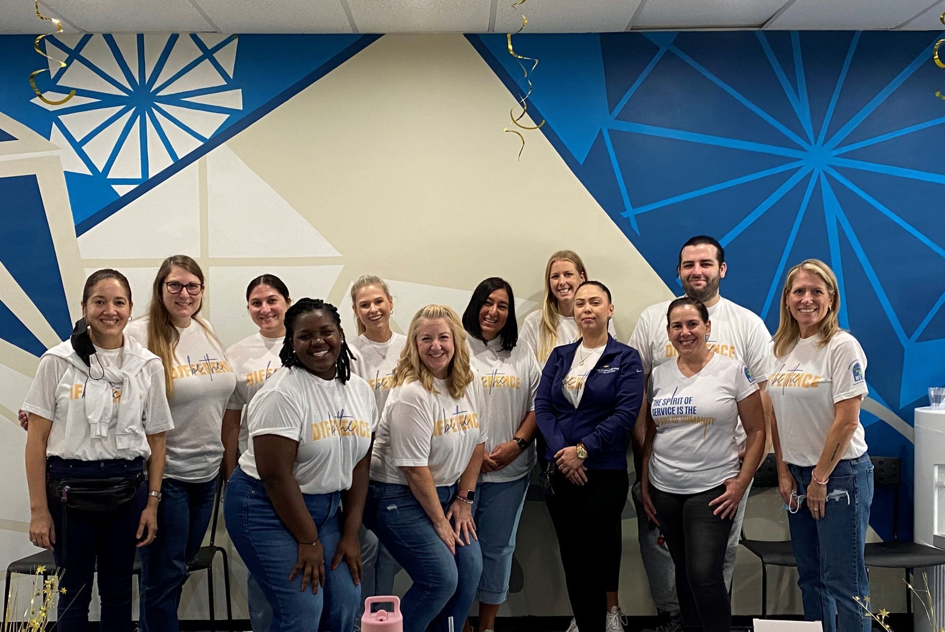 Group of people posing in front of a blue geometric mural, many wearing white shirts and jeans.