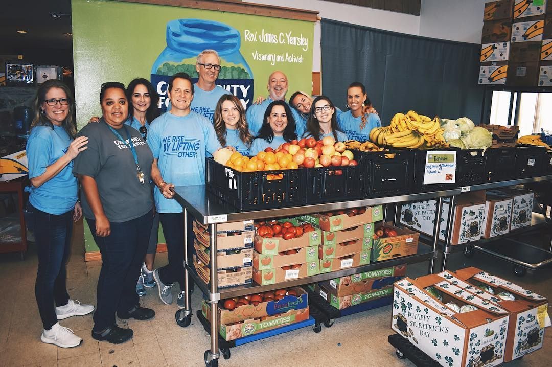 Group of volunteers standing behind tables of donated produce and canned goods at a community food drive.