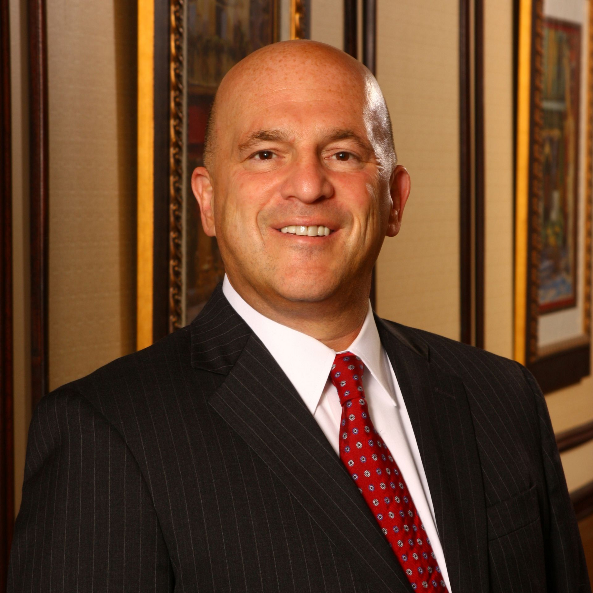 Smiling man in a dark suit with a red tie standing indoors near framed artwork