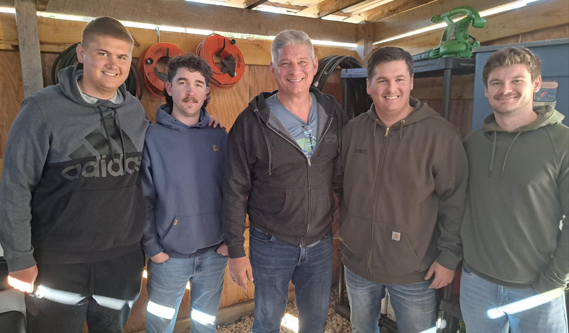 Five smiling men standing together in a rustic wooden shed, wearing casual jackets and hoodies.