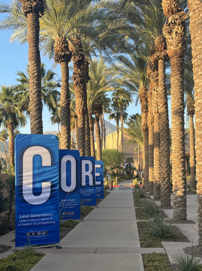 Palm-lined walkway with large blue “CORE” signboards along a sidewalk in bright sunlight