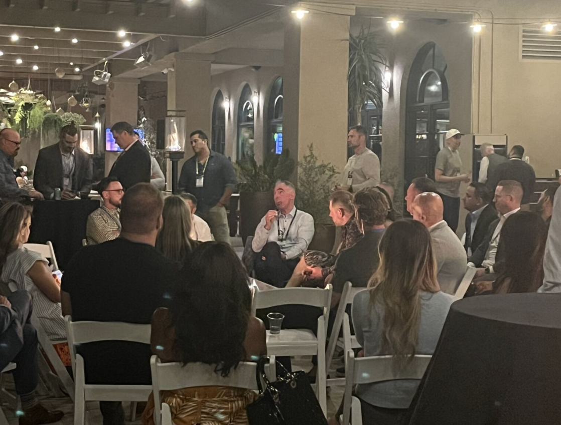 People seated in conversation at a dimly lit outdoor restaurant with arched windows and string lights.