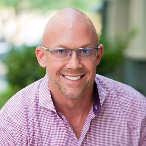 Smiling bald man in glasses wearing a pink checkered shirt, seated outdoors against a blurred green background