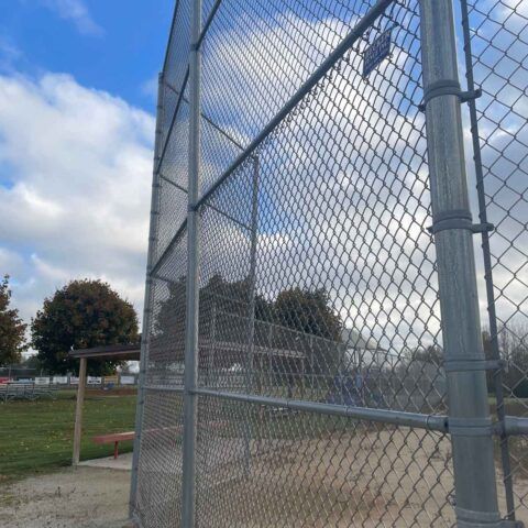 Chain-link baseball backstop beside a grassy field under a partly cloudy blue sky.