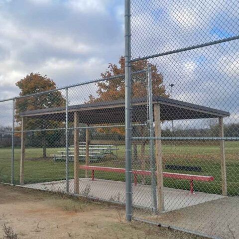 Covered baseball dugout behind a chain-link fence, with benches and a field in the background.