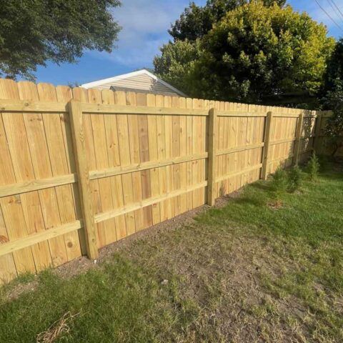 New wooden privacy fence along a grassy backyard, with trees and a house roof visible behind it.