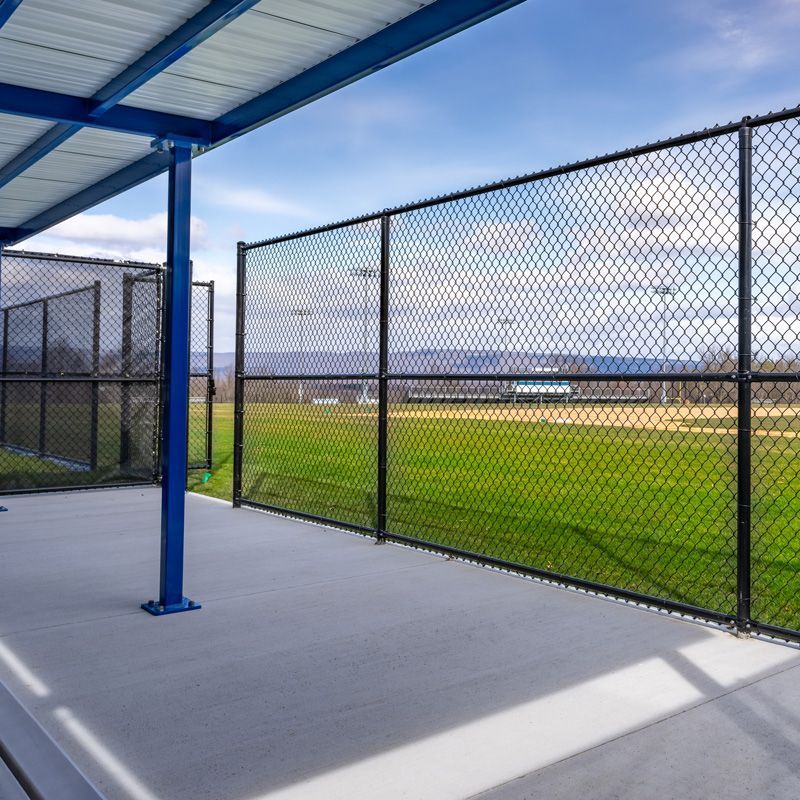 Covered concrete walkway beside a chain-link fence overlooking a grassy sports field under blue sky
