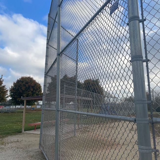 Chain-link fence beside a dirt baseball field under a partly cloudy sky
