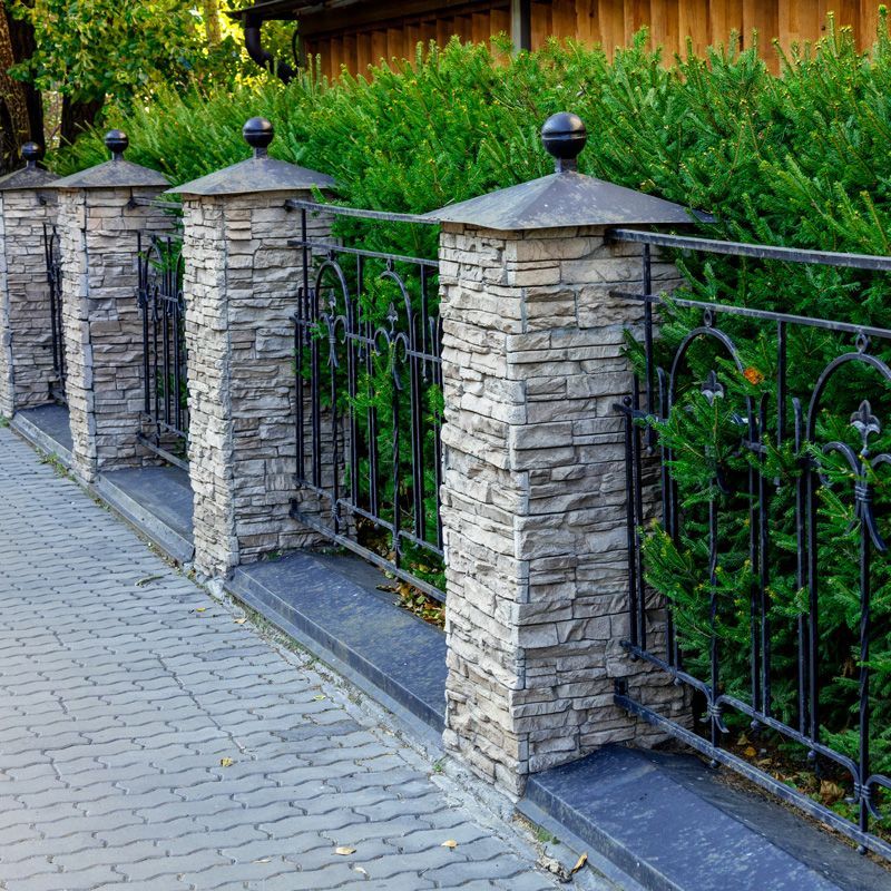 Stone fence with black metal railing along a paved sidewalk beside dense green hedges and a wooden building