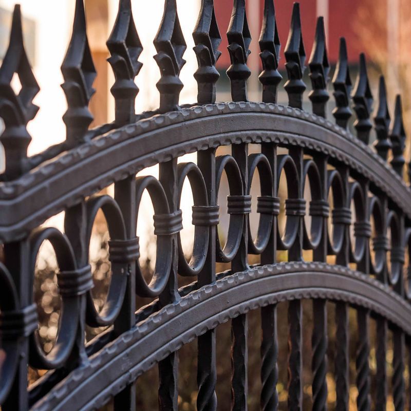 Black wrought-iron fence with ornate arched details and pointed spikes, close-up against a blurred background