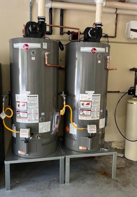Two gray water heaters on metal stands in a utility room, with pipes and valves above them