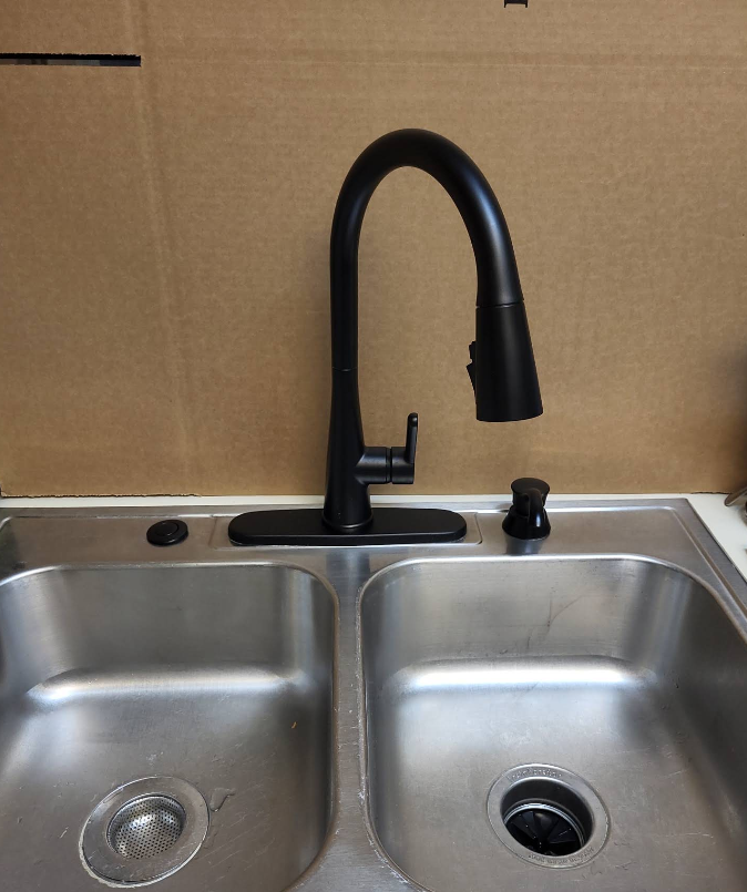 Stainless steel double sink with black gooseneck faucet against a cardboard backsplash.