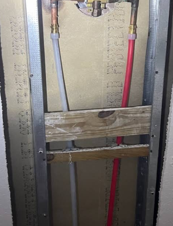 Interior wall framing with plumbing pipes, including red and gray lines, behind wooden braces.