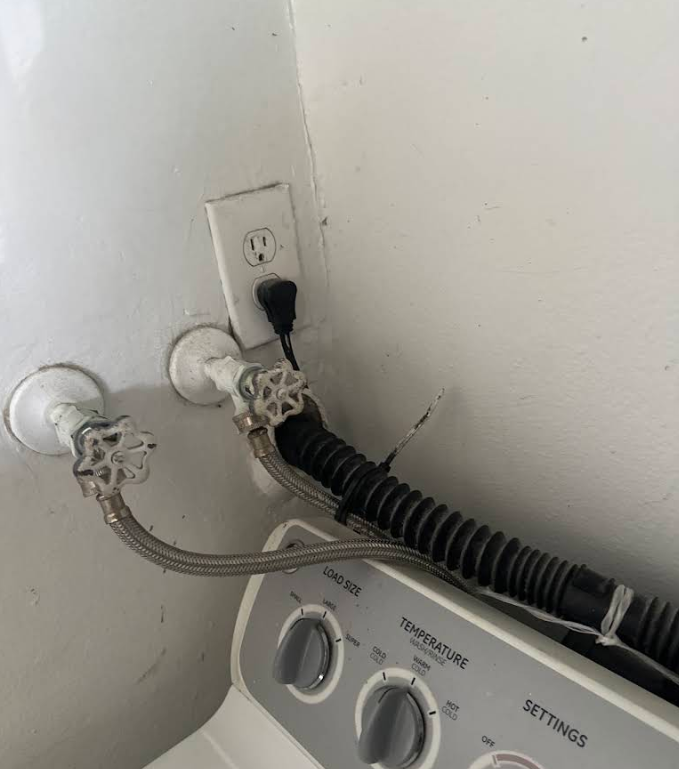Laundry washer hoses plugged into a wall outlet behind a washing machine in a corner.