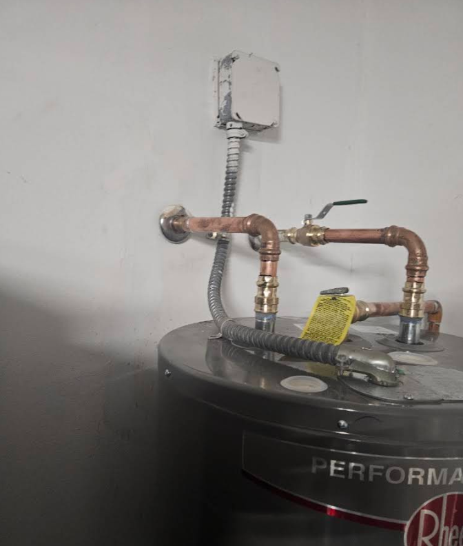 Exposed copper pipes and valves atop a water heater with a flexible conduit against a wall
